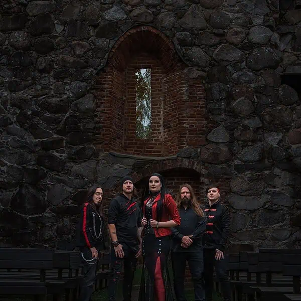 Five musicians in black and red outfits pose in a ruined stone courtyard with a brick-arched window behind them.