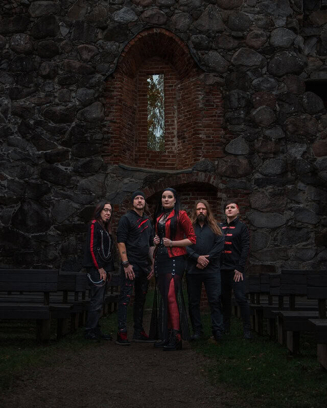Five musicians in black and red outfits pose in a ruined stone courtyard with a brick-arched window behind them.