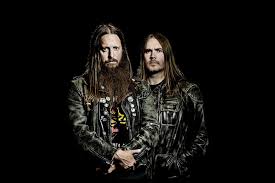 Two men with long hair wearing black leather jackets pose together against a dark studio backdrop.