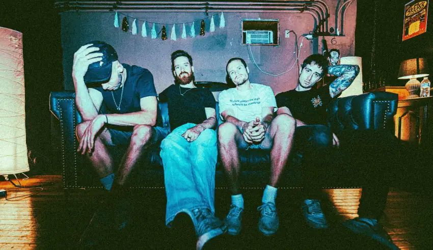 Four men sit on a dark couch in a dim room, casual clothes, one resting his head in his hand.