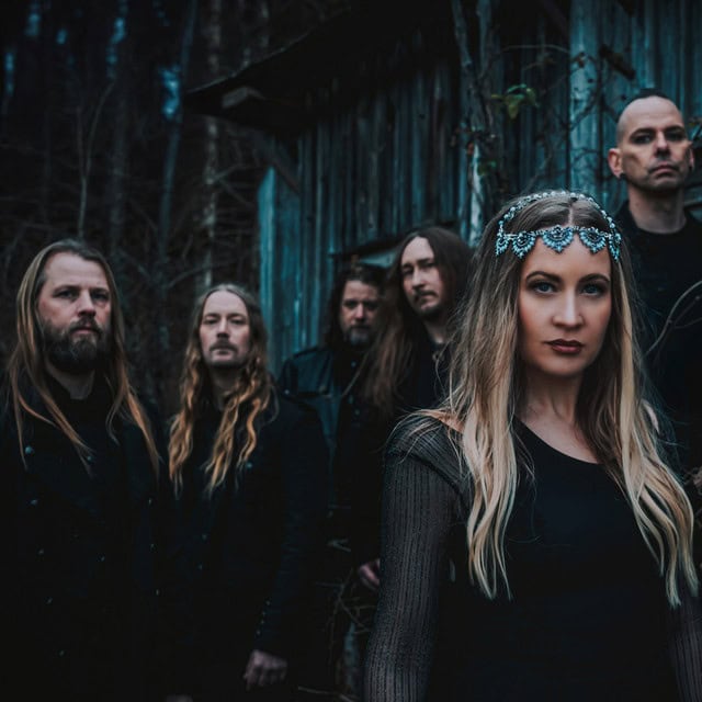 Frontwoman with a silver jeweled headpiece stands in the foreground, with a five‑member band in dark clothing behind her in a moody outdoor setting near a weathered wooden structure.