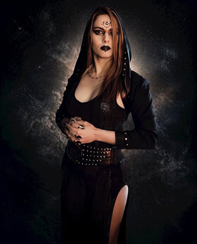 Gothic woman in a black hooded outfit with studded belt, dark makeup, and spacey backdrop.