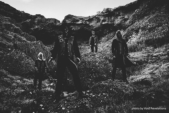 Black-and-white photo of four people standing on a rocky hillside in dark clothing, with a moody, windswept landscape. (photo by Void Revelations)