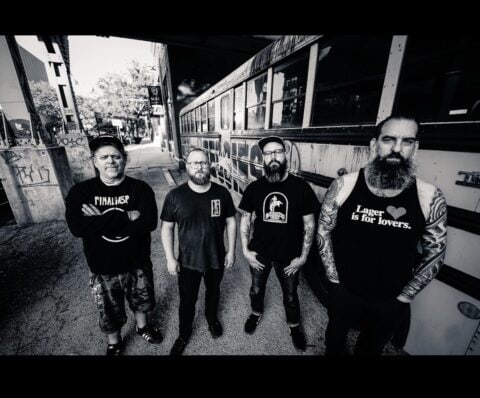 Four men in dark T-shirts stand in a street beside a bus, looking at the camera.