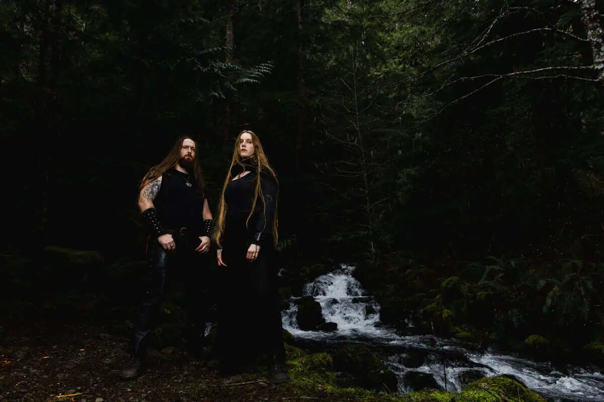 Two people with long hair in black clothing stand by a mossy forest stream.