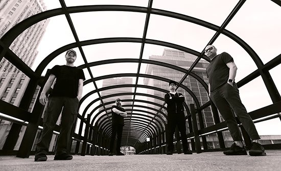Four men stand on a circular-arched pedestrian bridge in a city, posing for the camera.