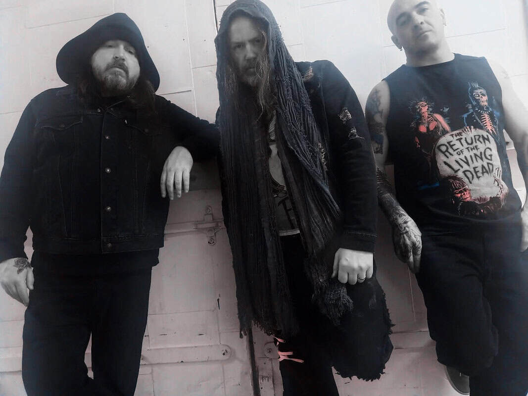 Three men in dark, edgy clothing lean against a white wall, one hooded, another with a tattered scarf, all with serious expressions.