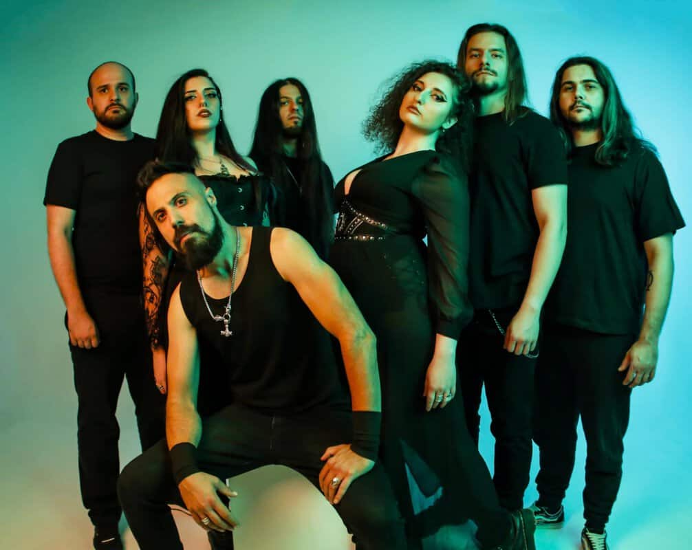 Six-member band in black outfits posing in a teal studio backdrop; front man kneels center with arms on his knees.