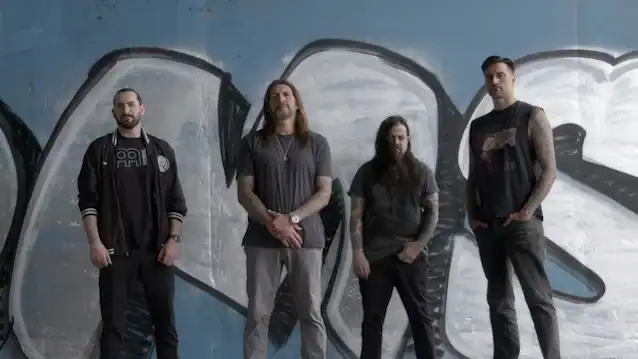 Four men stand in a row against a blue graffiti wall, posing like a rock band in casual dark clothing.