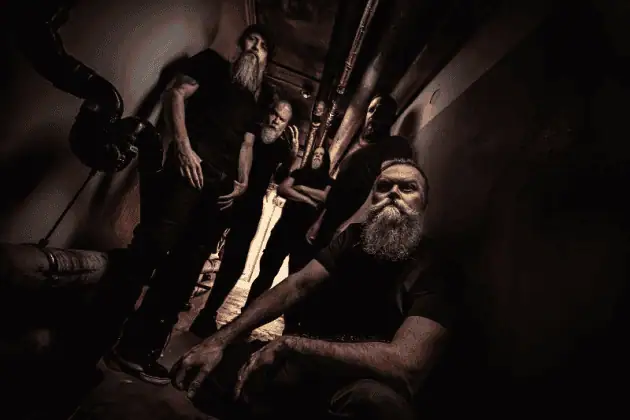 Bearded men in black clothing pose in a dark, industrial hallway with exposed piping behind them, one man seated in front.