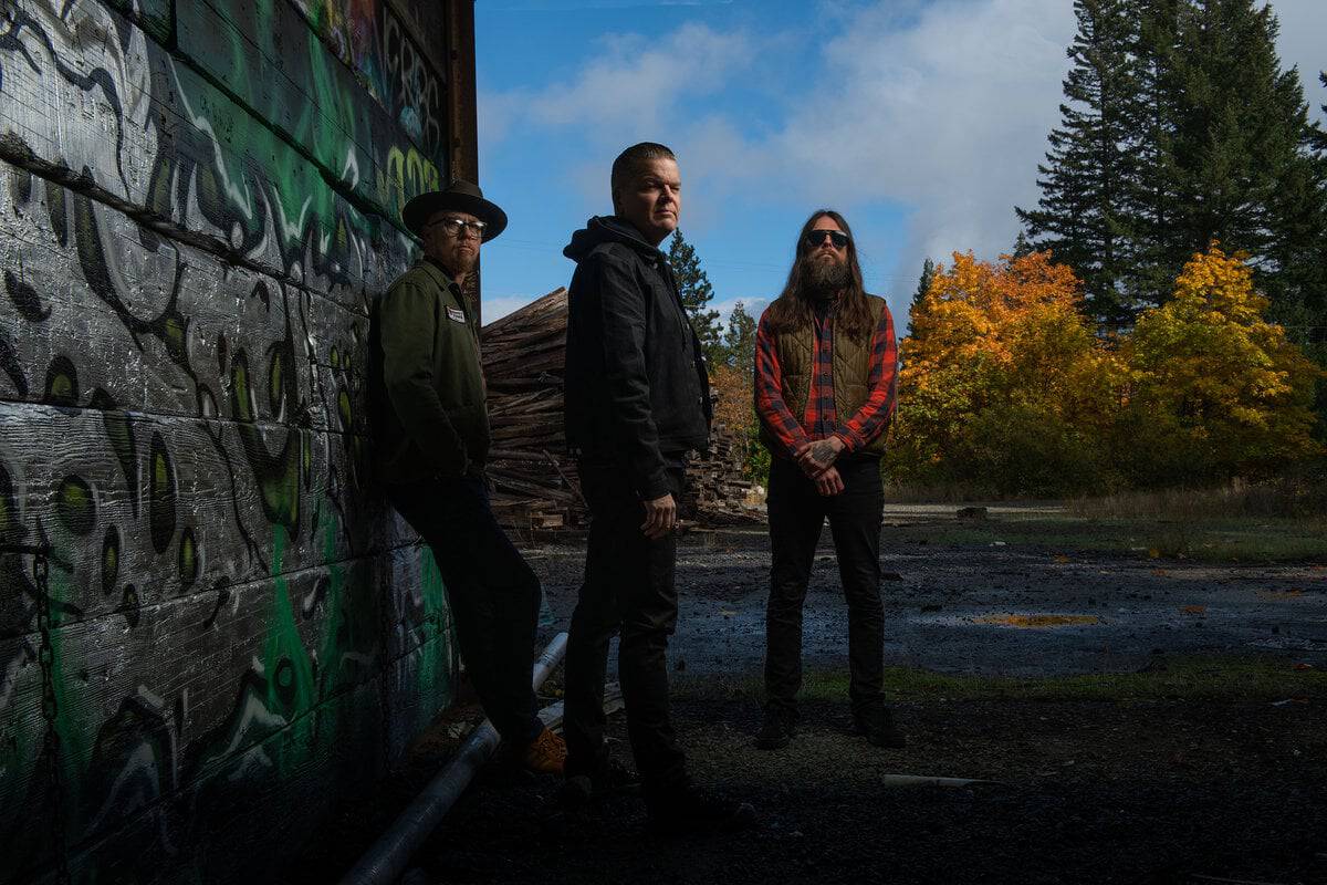 Three men stand beside a graffiti-covered wall in an outdoor setting on a sunny autumn day in the yard behind a wooden structure with fallen leaves nearby.",