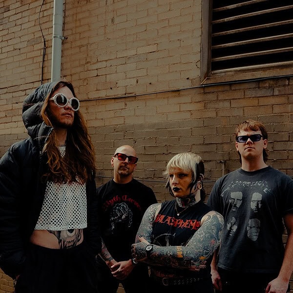Four rock/metal band members pose against a brick wall, wearing dark clothing and sunglasses, with tattoos and edgy hairstyles.