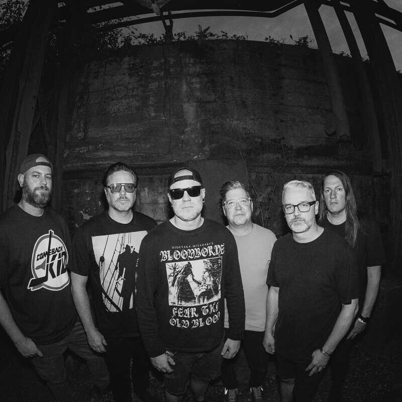 Six male rock band members pose for a group photo in a dark, industrial setting.