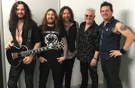 Five male rock band members pose together in a studio, wearing leather and denim with guitars and a coordinated rock look, smiling at the camera.