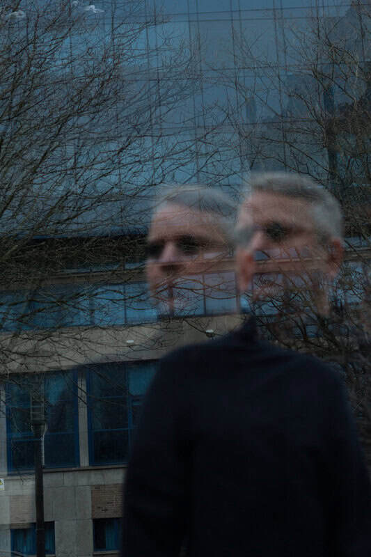 Man in a dark sweater reflected in a glass window, with bare tree branches and a blue building behind.
