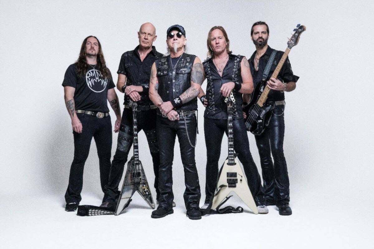 Five male rock musicians in black outfits pose with electric guitars in a white studio backdrop.