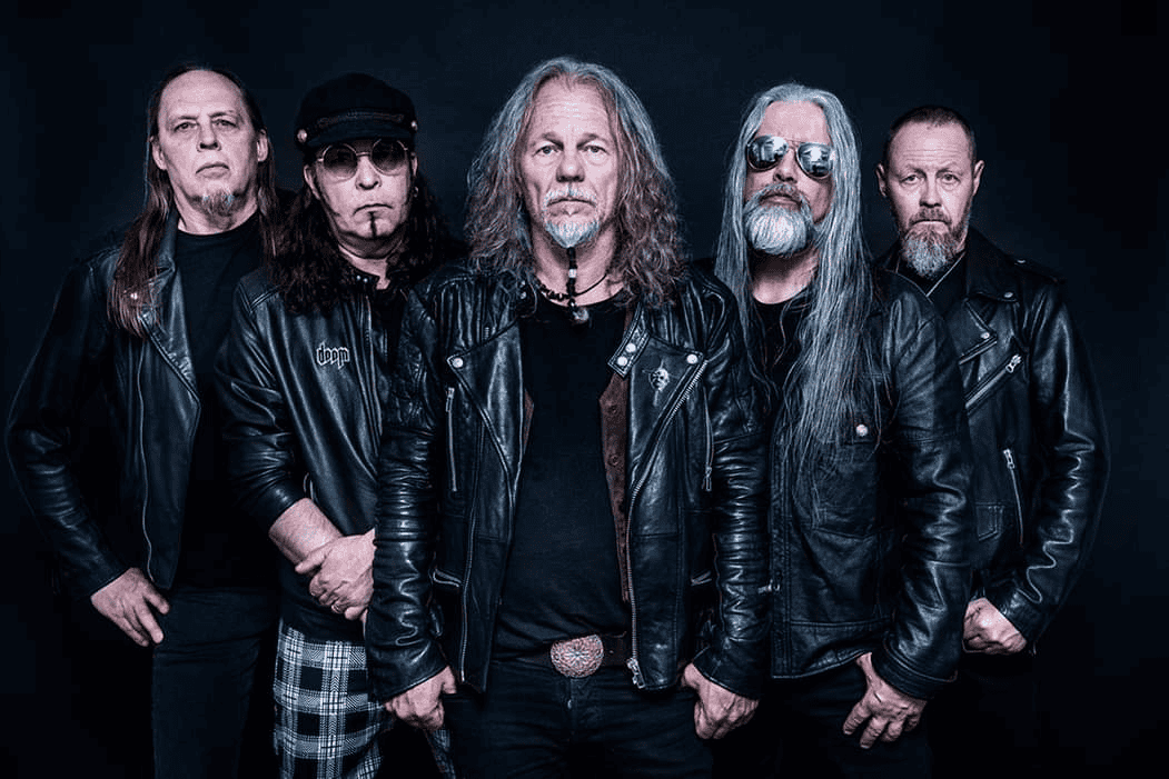 Five male rock musicians in black leather jackets posing together against a dark background.
