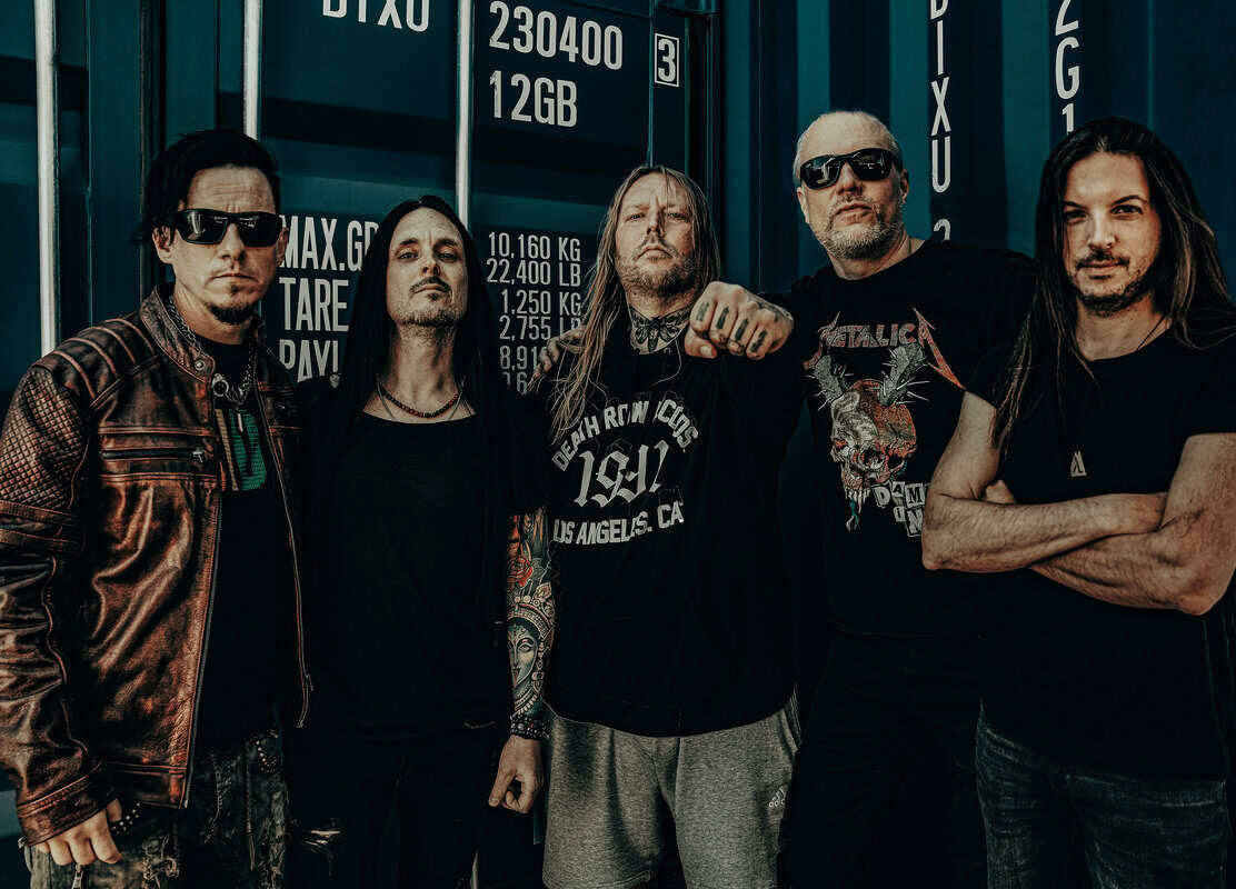 Five men in rock/metal attire pose together in front of blue shipping containers, some with tattoos and sunglasses, looking toward the camera.