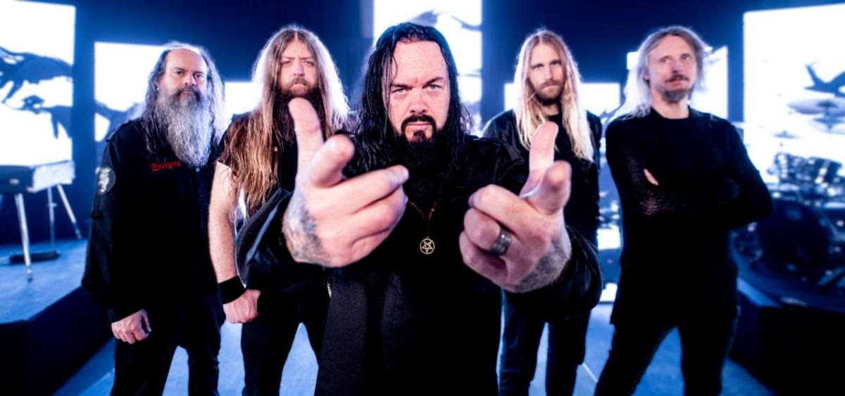 Five-member metal band posing for a promo shot, center member points at the camera with a serious expression and a pentagram necklace visible in front of a dark stage setup with bright blue lights behind them.