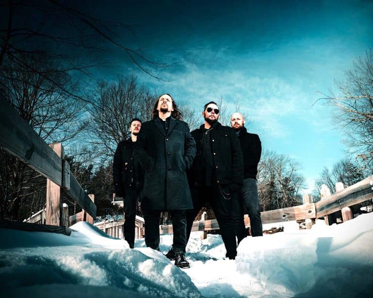 Four men in dark coats stand on a snowy bridge, posing for a band photo with a clear blue sky behind them.
