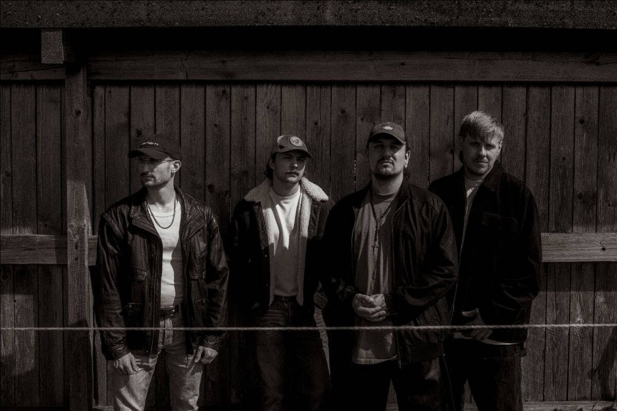 Four men in jackets stand against a weathered wooden fence, posing for a moody, black-and-white photo.