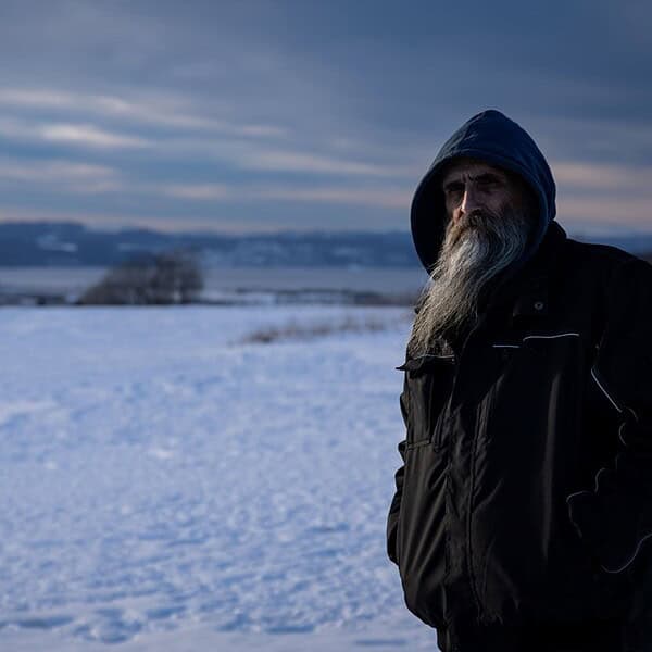 Bearded man in a blue hooded jacket stands in a snowy landscape at dusk with distant hills.
