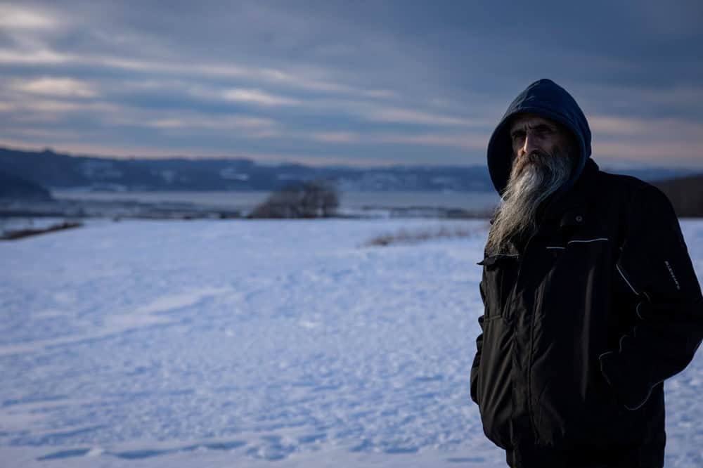 Bearded man in a blue hooded jacket stands in a snowy landscape at dusk with distant hills.