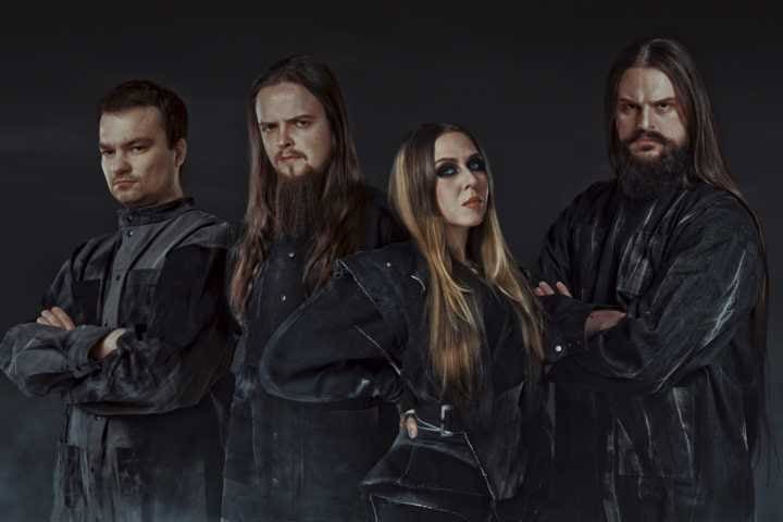 Four band members stand in black outfits against a dark background, two men with long hair and a woman with heavy makeup, posing confidently.