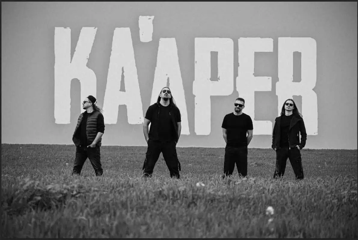 Four men in black stand in a field, sunglasses on, in front of a wall with large white letters behind them.