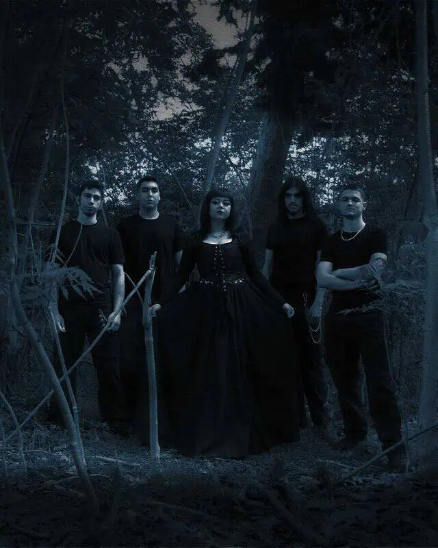 Five people in a dark forest posing for a gothic-themed photo; a woman in a long black corseted gown stands center, flanked by four men in black shirts, some holding prop weapons.