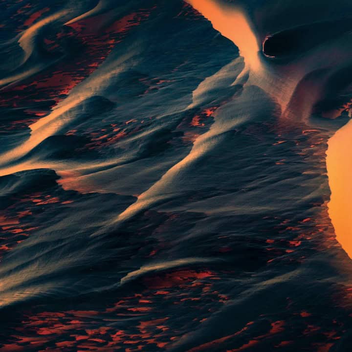 Abstract aerial view of wind-sculpted dunes in blue and orange hues at sunset, with flowing ridges and dramatic light.
