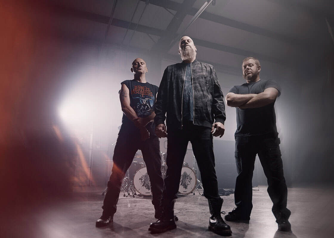 Three men stand in a dim rehearsal space with a drum kit behind them and orange halo lighting on the left.