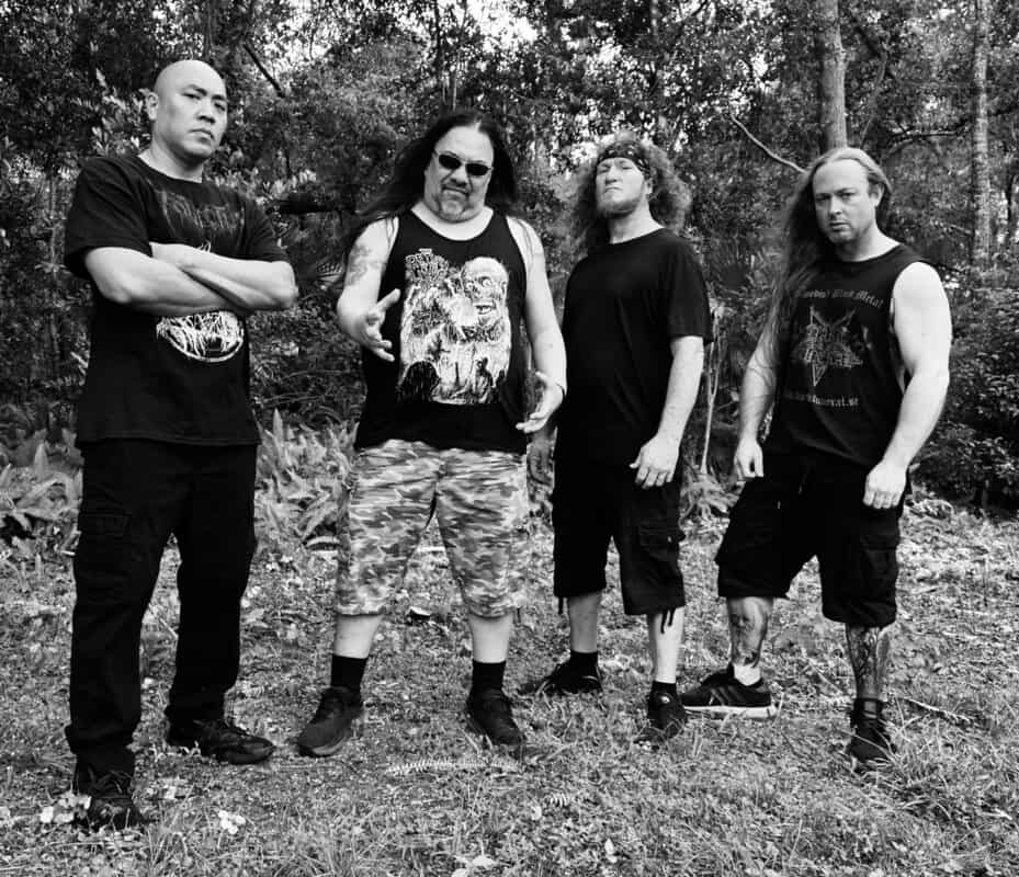 Four men wearing dark clothing pose outdoors in a forested area, creating a metal-band style group photo (black and white).