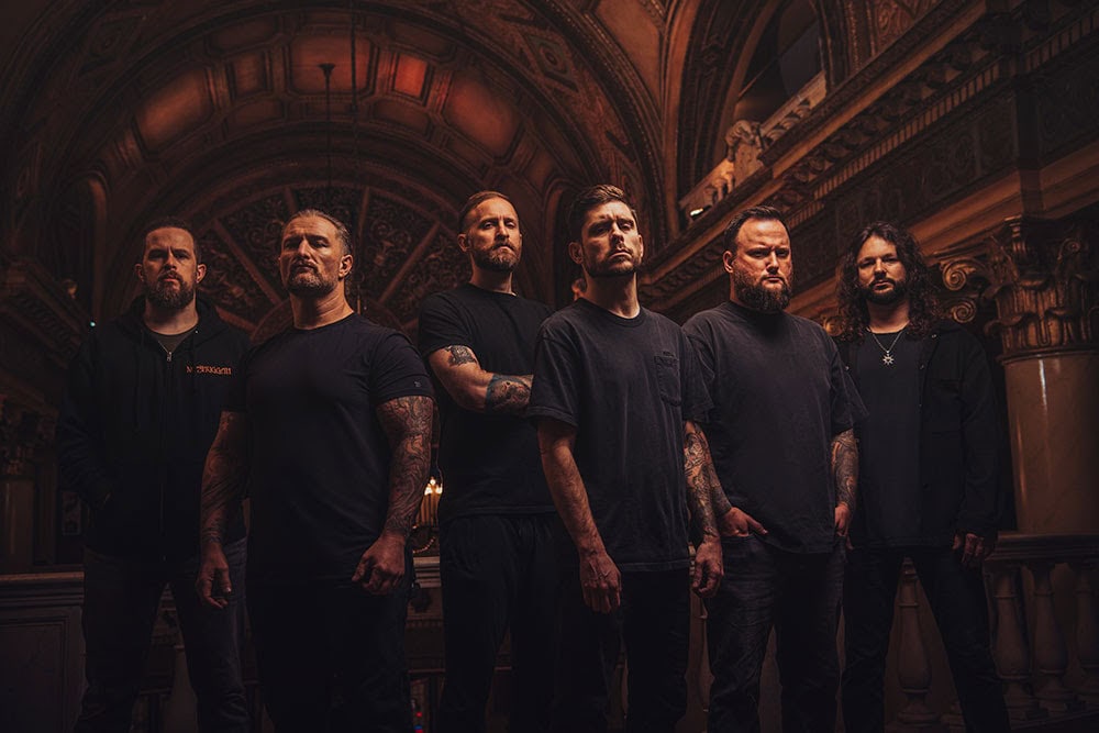 Six men with tattooed arms stand in a dim, ornate hall, facing the camera.