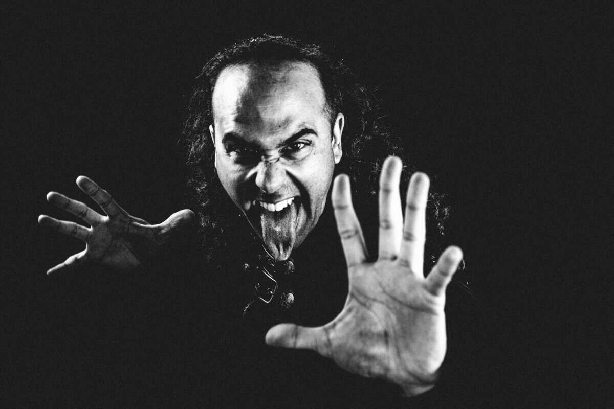 Black-and-white close-up of a man with long hair, grinning fiercely as both hands are pressed toward the camera, creating a dramatic, energetic portrait.