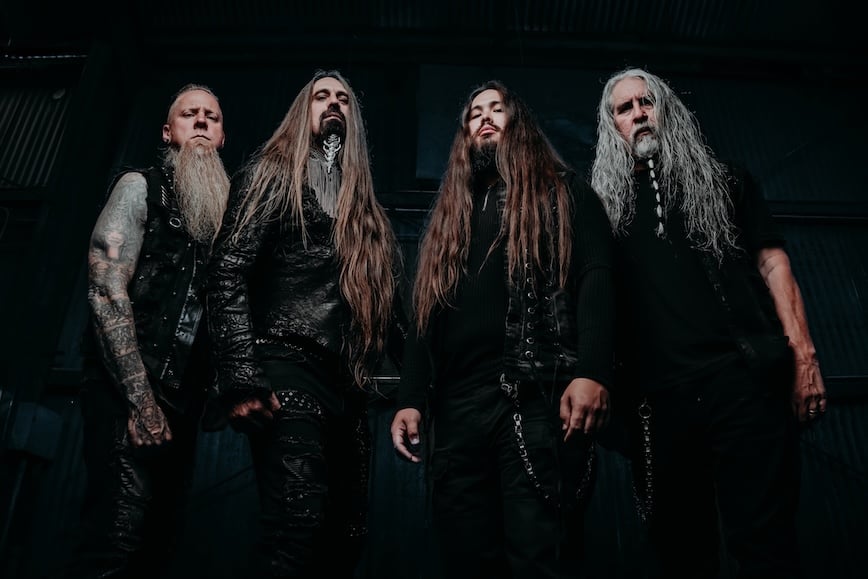 Four long-haired men in black metal attire standing in a dark industrial setting, facing the camera with serious expressions.