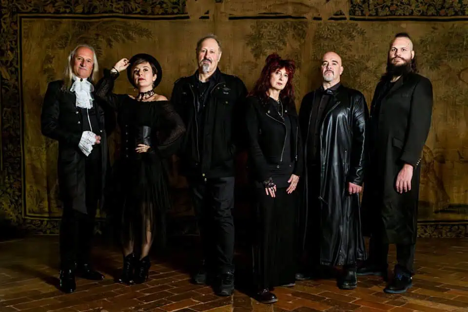 Six adults in black Gothic attire stand in a row indoors against a tapestry wall and wooden floor. A diverse band or group portrait.