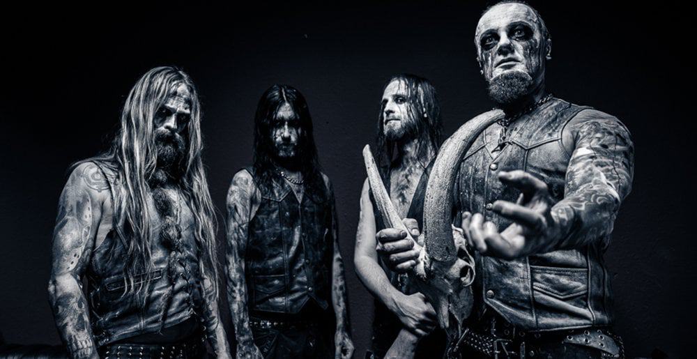 Four tattooed men in black leather outfits pose in a dark studio, creating a heavy-metal band portrait.