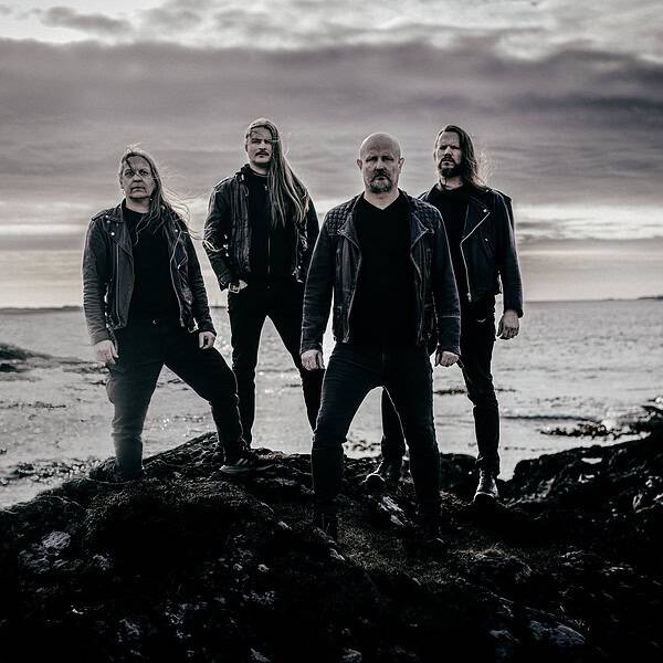 Four men in black leather jackets stand on a rocky shoreline under a cloudy sky, facing the camera.