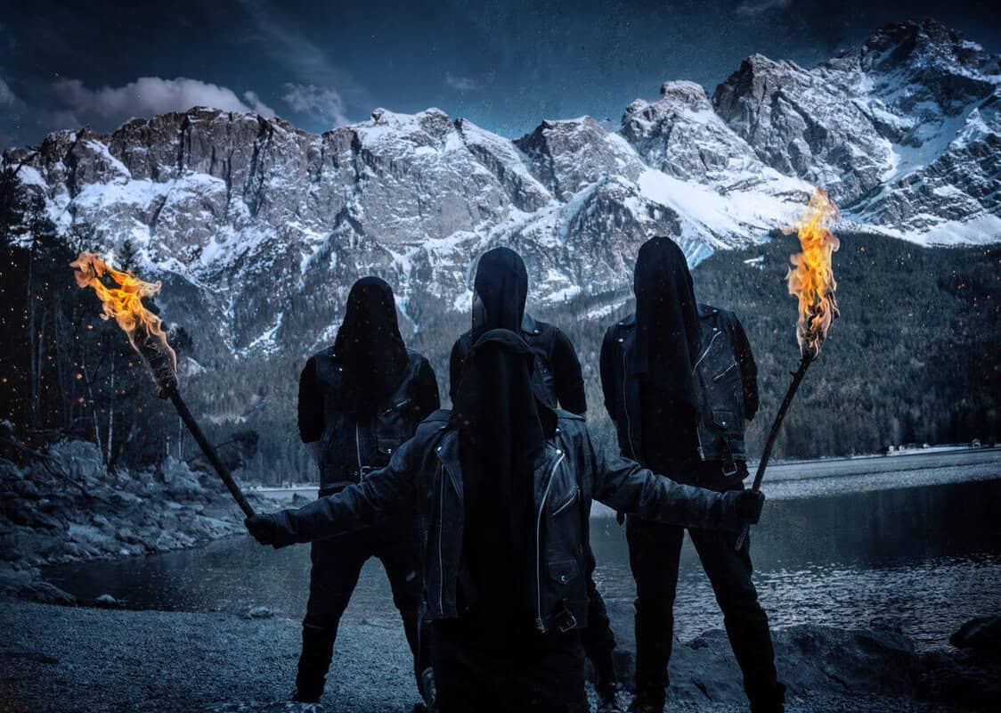 Three hooded figures stand by a snowy lake at night, holding blazing torches as mountains loom behind them.