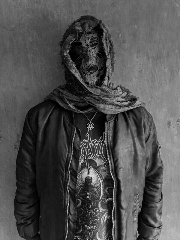 Person wearing a torn hood and skeletal mask over the face, in a dark jacket, standing against a concrete wall.