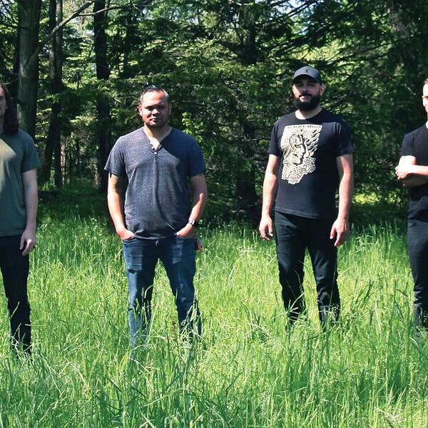 Five men stand in a grassy field with a forest backdrop, facing the camera in casual shirts and jeans.