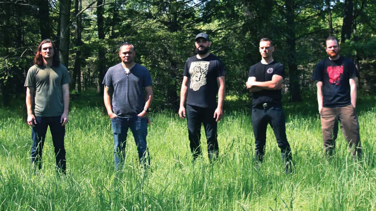 Five men stand in a grassy field with a forest backdrop, facing the camera in casual shirts and jeans.