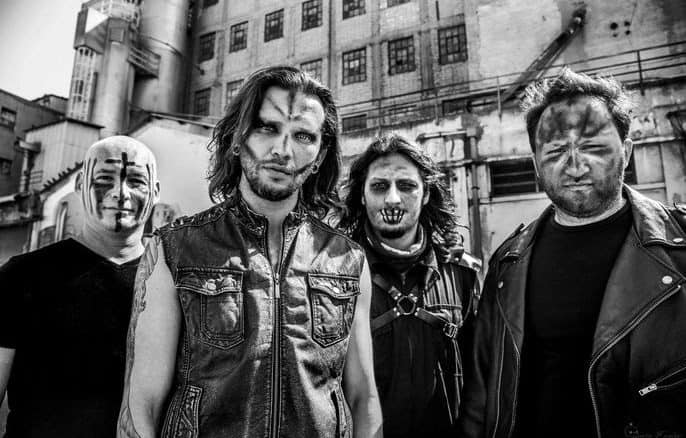 Four male heavy metal musicians with face paint pose in a gritty industrial setting, wearing leather jackets in a black-and-white photo.