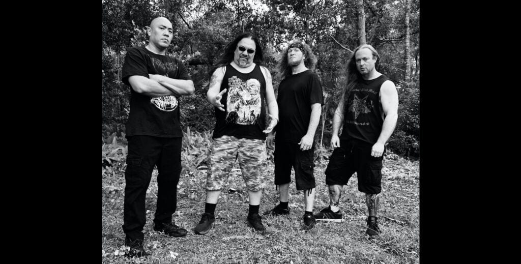 Four men stand together in a wooded outdoor setting, dressed in dark casual attire for a rock/metal band pose (black-and-white photo).