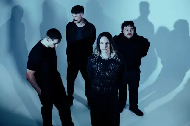 Five men in dark clothing pose in a blue-lit studio, forming a moody band portrait.