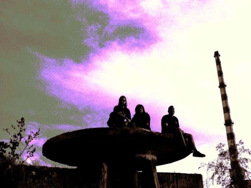 Three people sit on a large round concrete platform outdoors, with a tall striped smokestack to the right under a purple-tinged sky