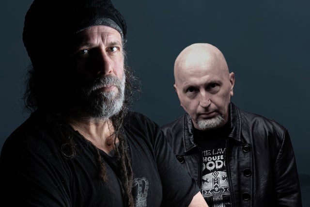 Two men in a studio portrait: one with a beanie and gray beard, the other bald in a black leather jacket against a dark backdrop.