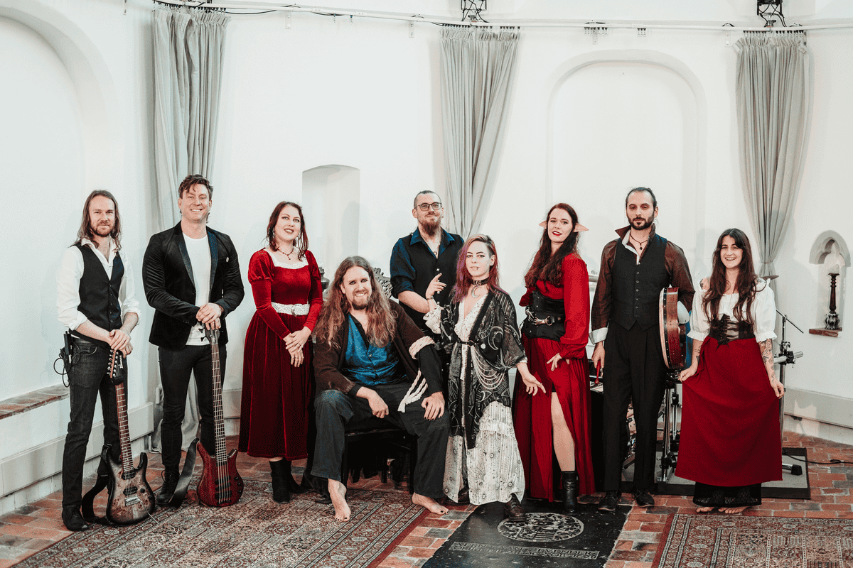 Ten musicians in period costumes pose with instruments in a bright rehearsal room with curtains.