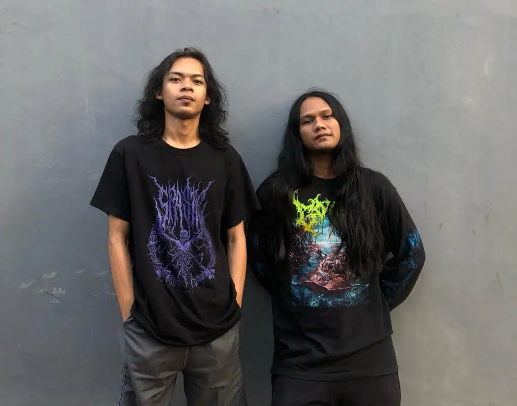 Two people with long hair stand against a gray wall, wearing black graphic band shirts with colorful logos.